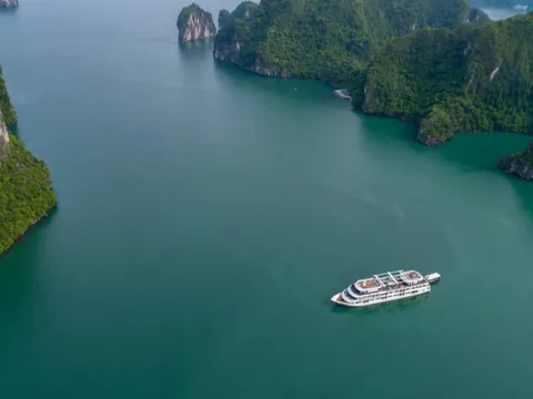 Aerial view of a white cruise ship sailing through emerald green water surrounded by tall limestone karst islands covered in vegetation.