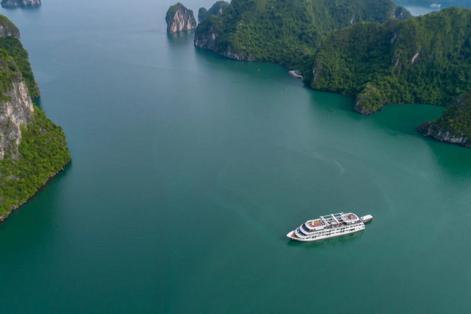 Aerial view of a white cruise ship sailing through emerald green water surrounded by tall limestone karst islands covered in vegetation.
