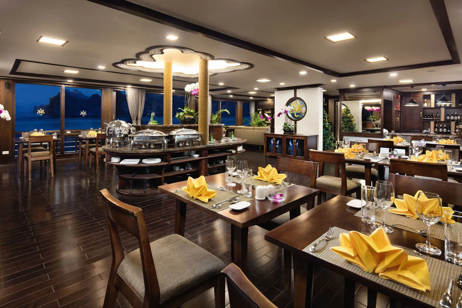 Interior of a dark-wood restaurant with set tables, yellow folded napkins, a buffet counter with chafing dishes, and night windows showing limestone karst silhouettes.