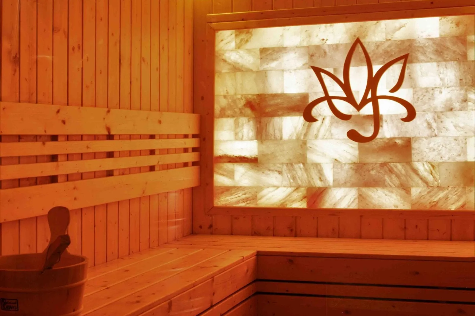 Interior of a wooden sauna with tiered benches, a bucket and ladle, a slatted wall lamp, and a backlit salt-brick panel with a cut-out lotus-style emblem.