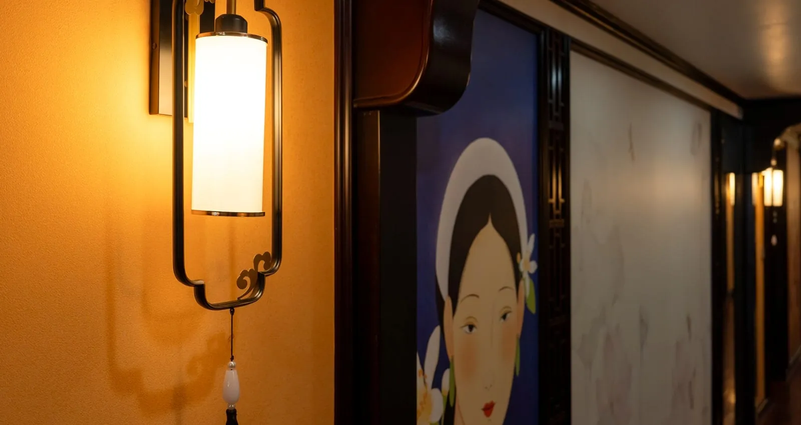 An indoor corridor with a lit wall sconce on an orange wall, a painted portrait of a woman in a white headdress with flowers, and a polished wooden floor leading to cabin doors.