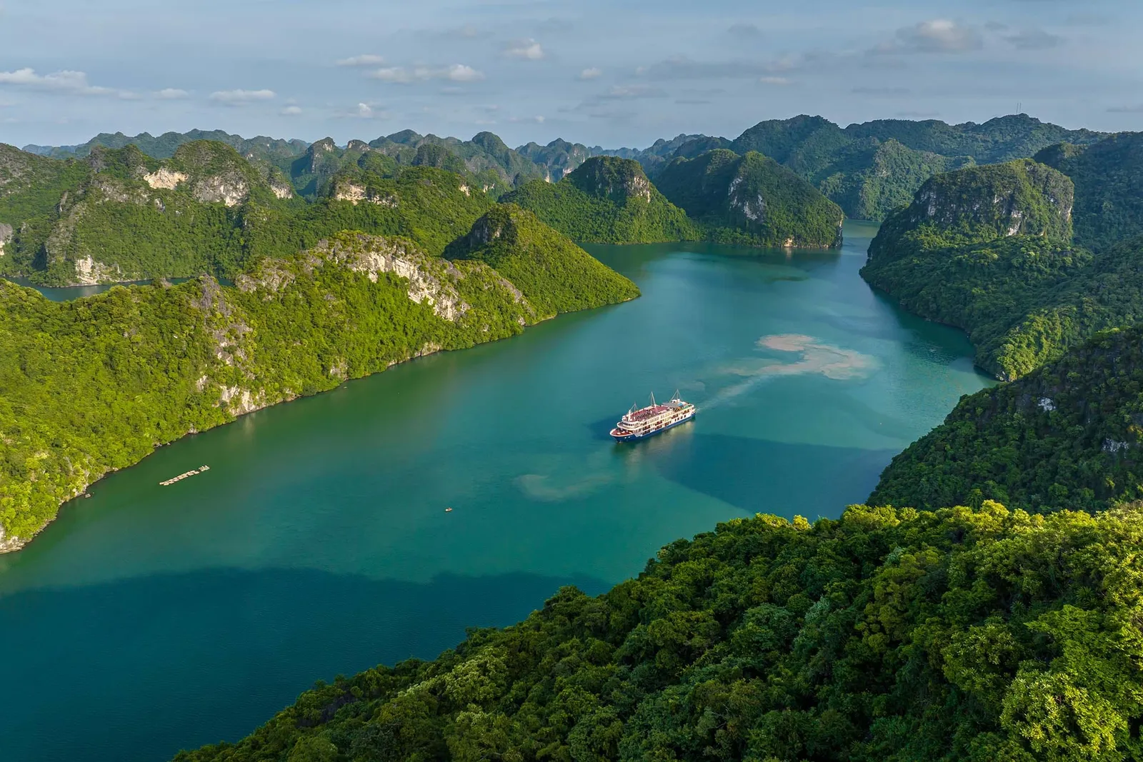 An elevated aerial view of a single cruise boat on turquoise water, surrounded by steep green limestone karst islands covered in forest, with a floating platform near the left shore.