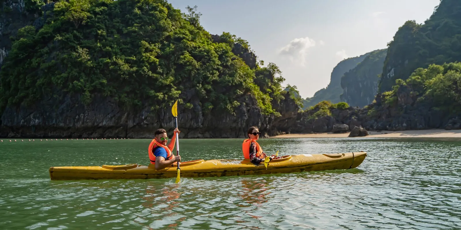 Two people in orange life jackets paddle a yellow tandem kayak on green water beside forested limestone karst cliffs and a small sandy beach.