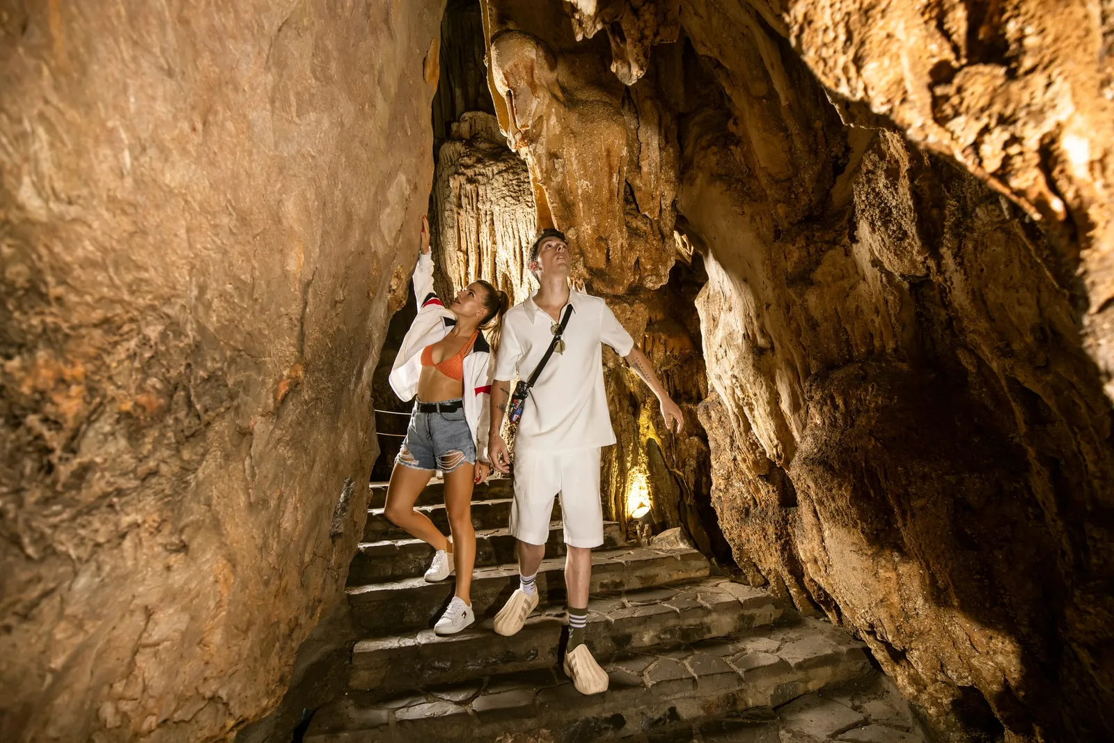 Two tourists standing on stone steps inside a lit cave with stalactites and rock formations.