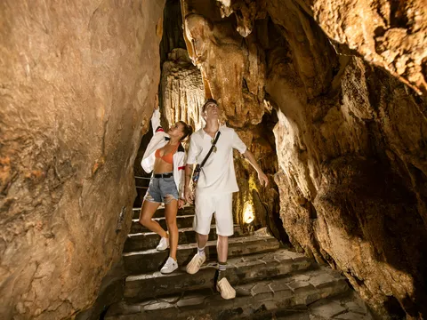 Two tourists standing on stone steps inside a lit cave with stalactites and rock formations.