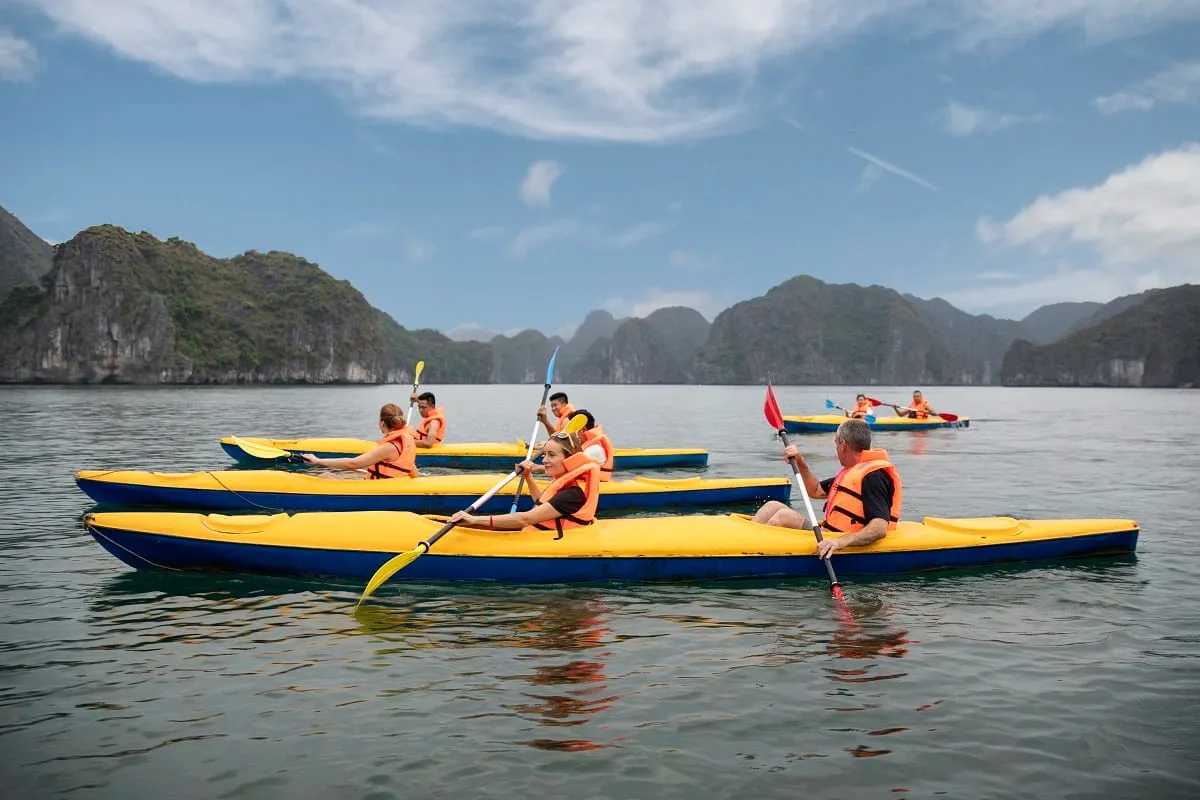 People wearing orange life vests paddle yellow and blue kayaks on calm water with limestone karst mountains in the background.