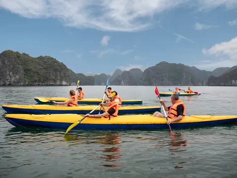 People wearing orange life vests paddle yellow and blue kayaks on calm water with limestone karst mountains in the background.