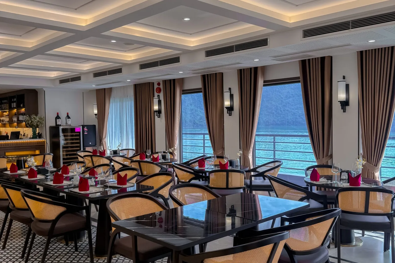 Interior of a ship's dining room with black tables, rattan-back chairs, red napkins, patterned tile floor and large windows showing turquoise water and forested limestone hills.