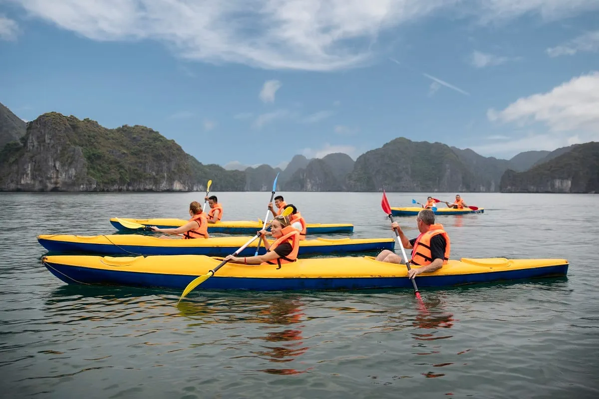 Several people wearing orange life jackets paddle yellow and blue kayaks on calm water with limestone karst formations in the background.