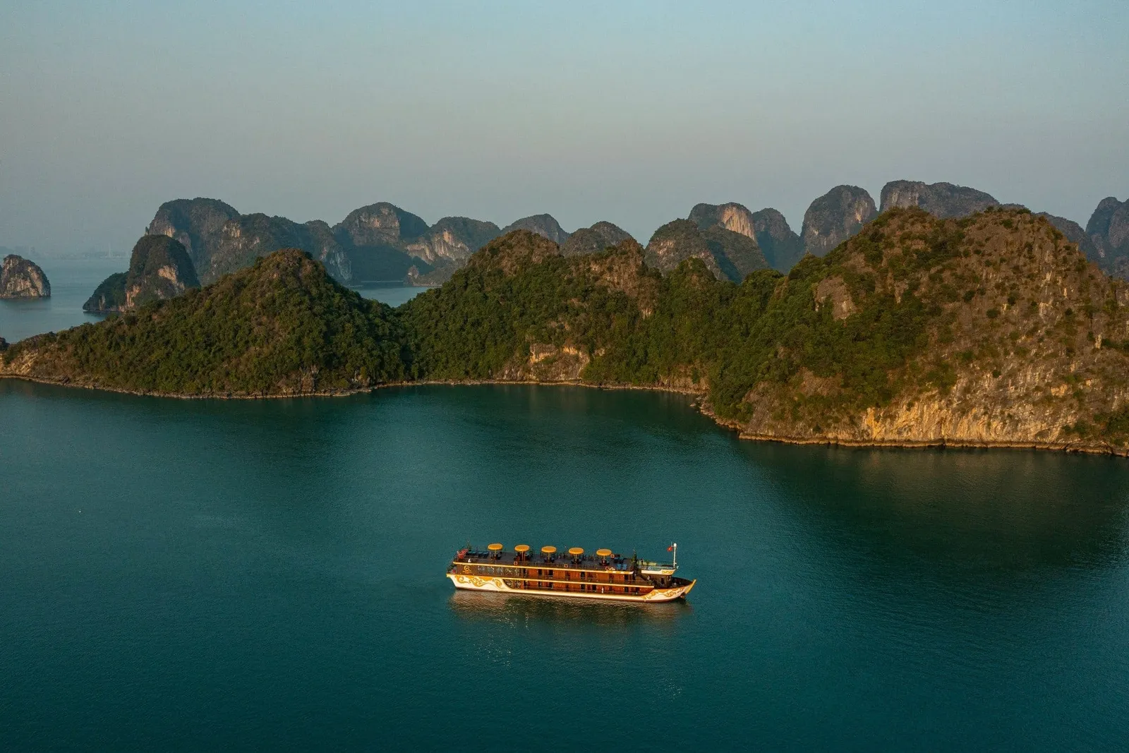 Aerial view of a small multi-deck brown and cream vessel with round yellow parasols on its top deck, sailing on calm teal water in front of forested limestone karst islands at dusk.