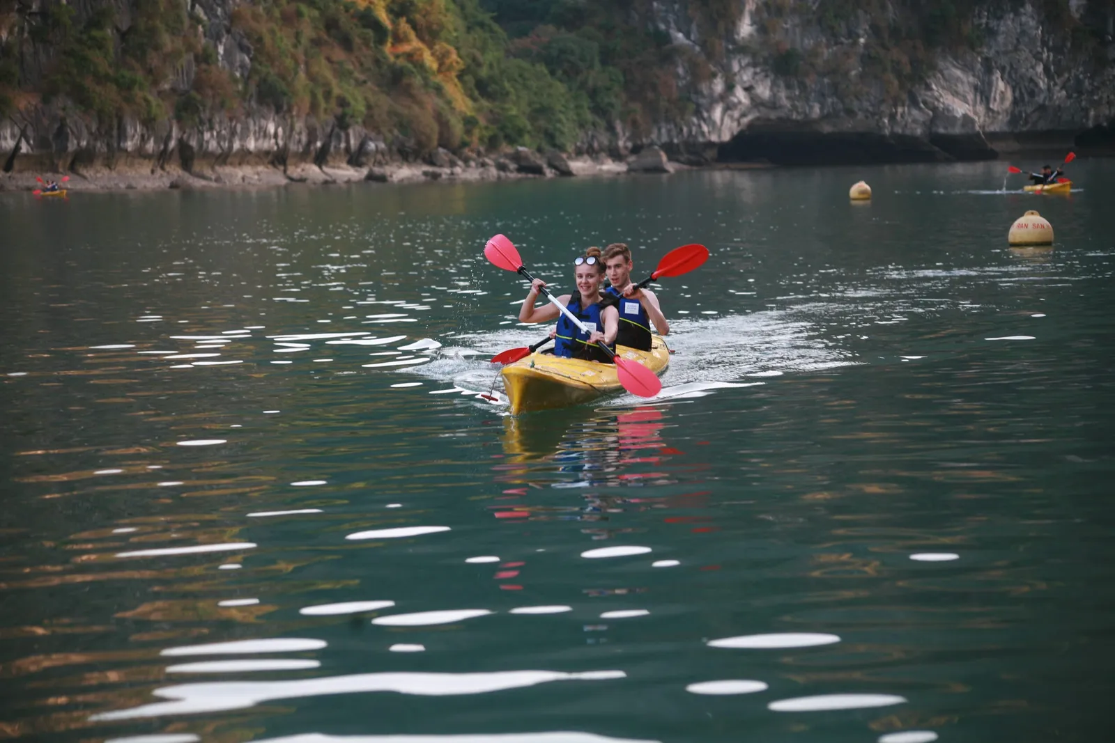 Two people in life jackets paddle a yellow kayak on calm green water with limestone karst cliffs in the background.