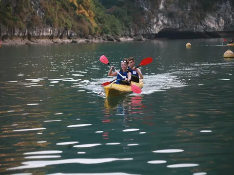 Two people in life jackets paddle a yellow kayak on calm green water with limestone karst cliffs in the background.