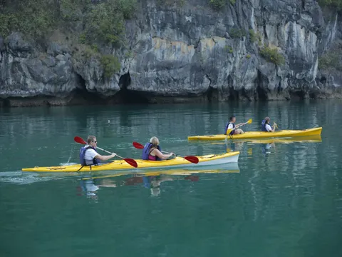 Four people kayaking in yellow kayaks on calm green water in front of a tall limestone karst cliff.