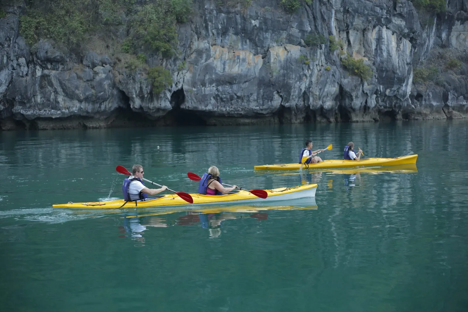Four people kayaking in yellow kayaks on calm green water in front of a tall limestone karst cliff.