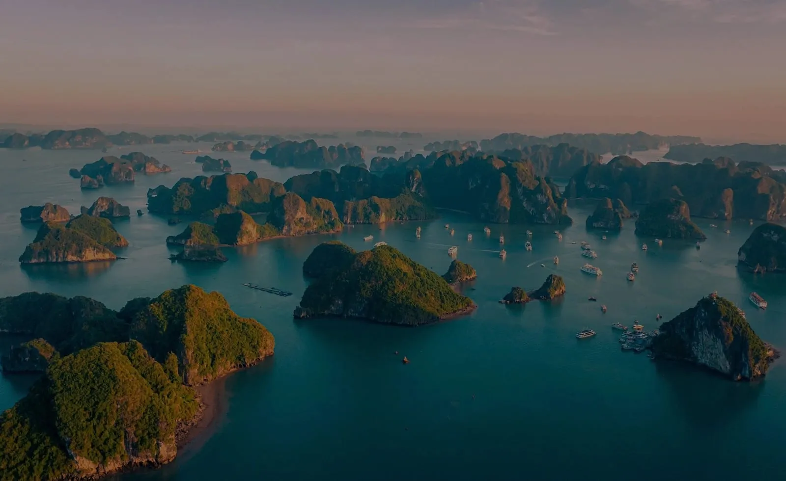 An aerial view at sunset showing dozens of limestone karst islands rising from turquoise water with small boats scattered between them.