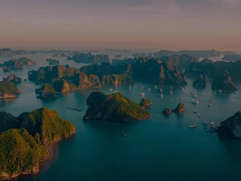 An aerial view at sunset showing dozens of limestone karst islands rising from turquoise water with small boats scattered between them.