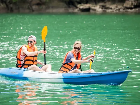 Two people wearing orange life jackets paddle a blue kayak on calm green water with a rocky shoreline behind them.