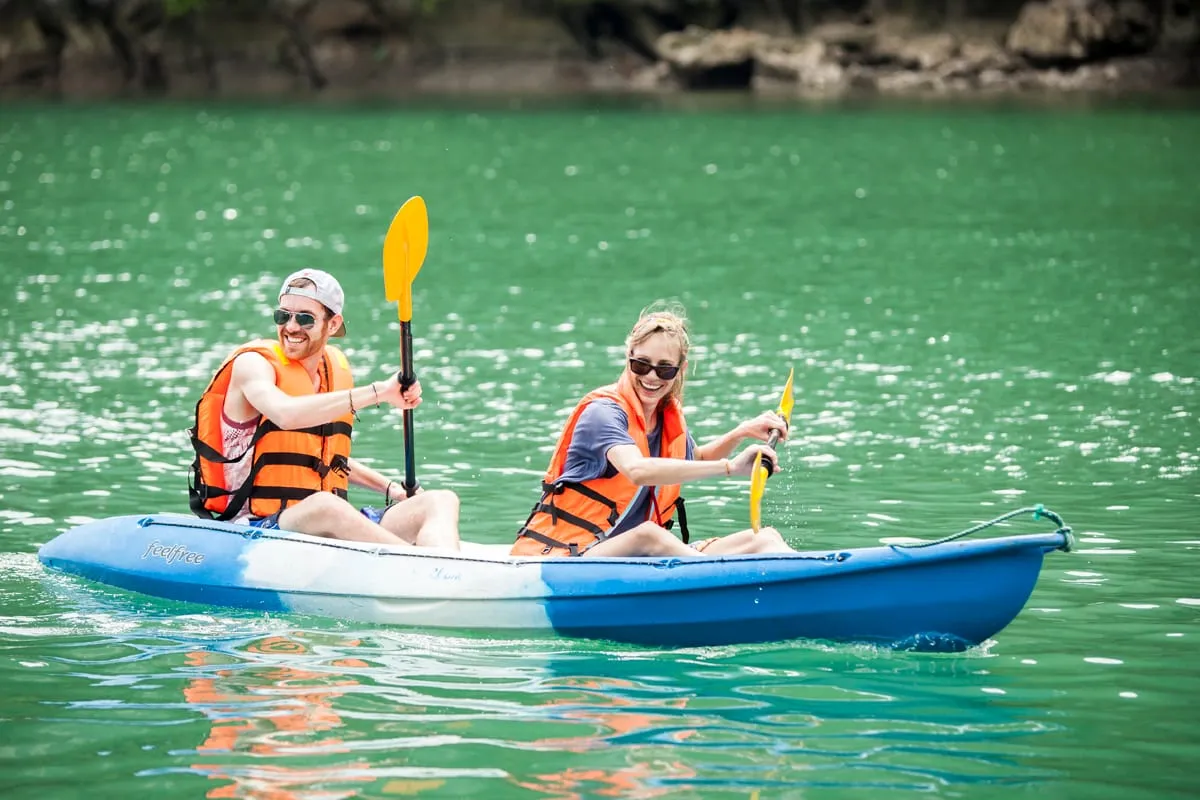 Two people wearing orange life jackets paddle a blue kayak on calm green water with a rocky shoreline behind them.