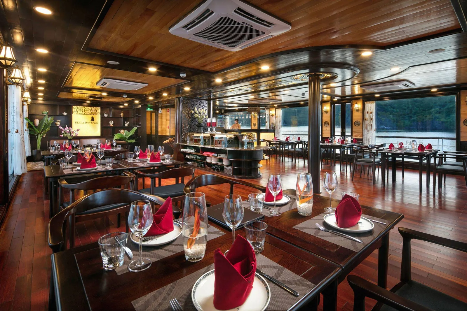 A dark-wood dining room with tables set with red folded napkins, wine glasses and plates, a buffet station with a staff member at the back, and windows showing a forested hillside over water, with a lit sign on the back wall reading 'Perla Dawn'.