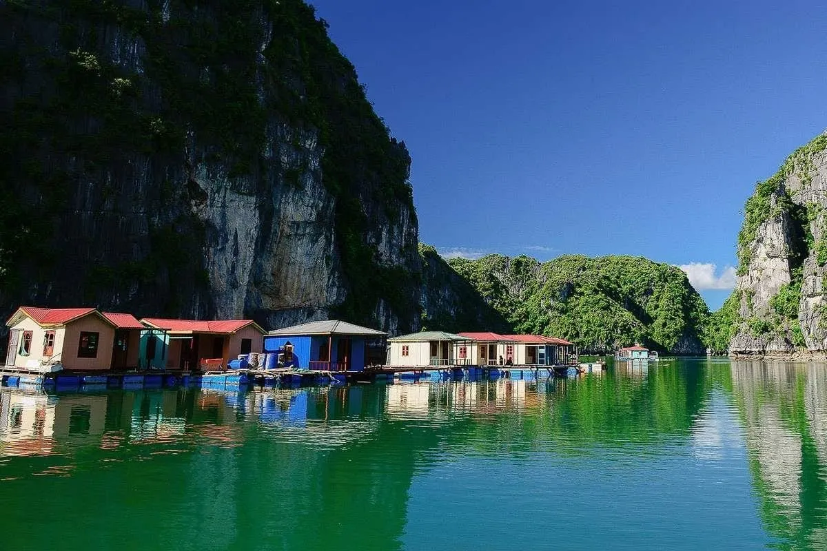 A row of colorful floating houses on blue pontoons beside steep green limestone cliffs, reflected in calm green water under a blue sky.