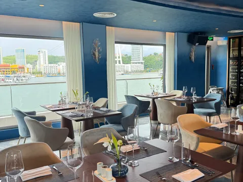 A blue-toned dining room with set tables, velvet chairs and a wine cabinet, with large windows looking out to the water, a waterfront of high-rise buildings, green hills and moored cruise boats.