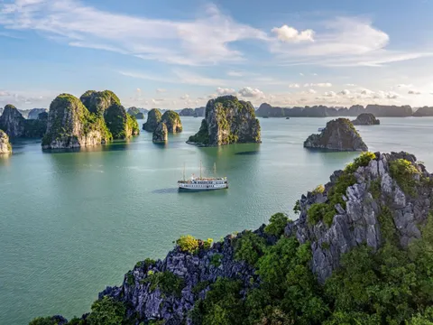 An aerial view of Ha Long Bay showing a white multi-deck cruise ship surrounded by numerous karst limestone islands rising from calm turquoise water under a partly cloudy sky.