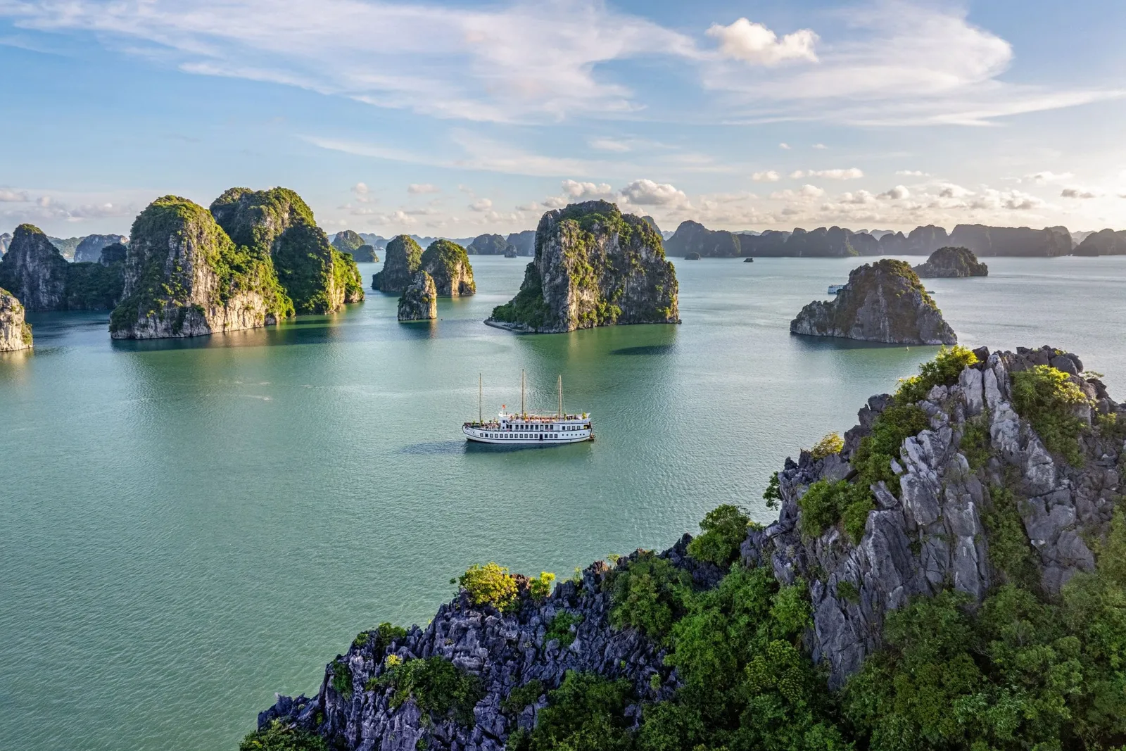 An aerial view of Ha Long Bay showing a white multi-deck cruise ship surrounded by numerous karst limestone islands rising from calm turquoise water under a partly cloudy sky.