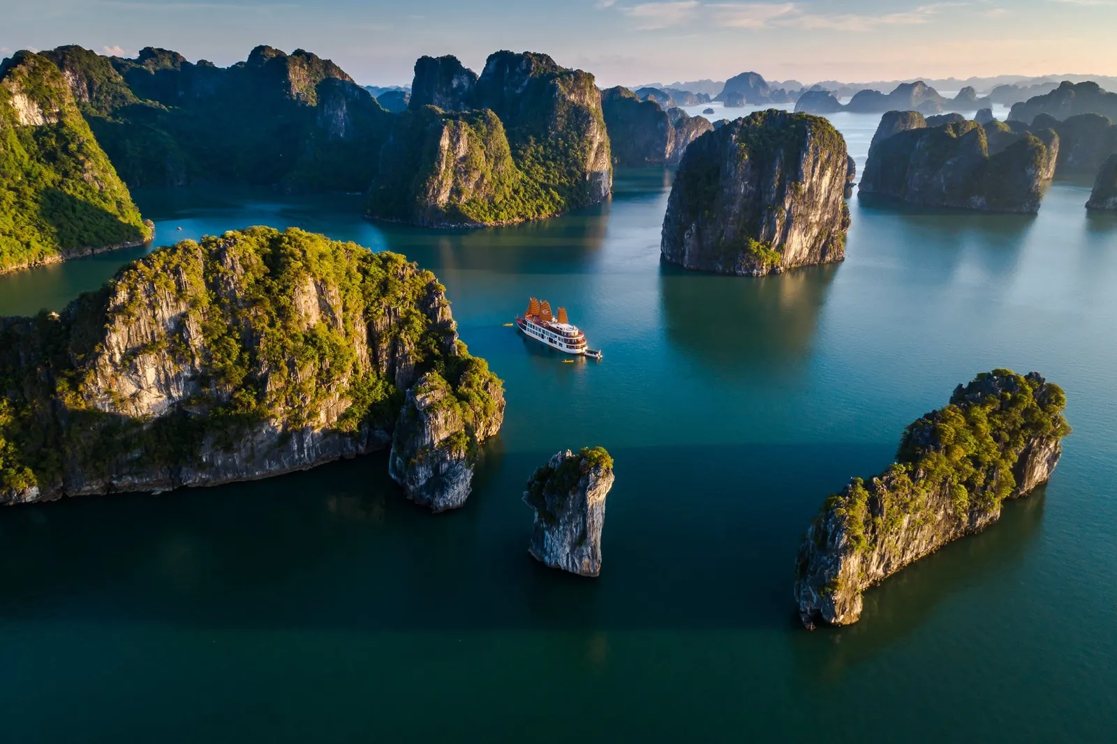Aerial view of a cruise boat with red sails navigating between tall limestone karst islands in calm turquoise water.
