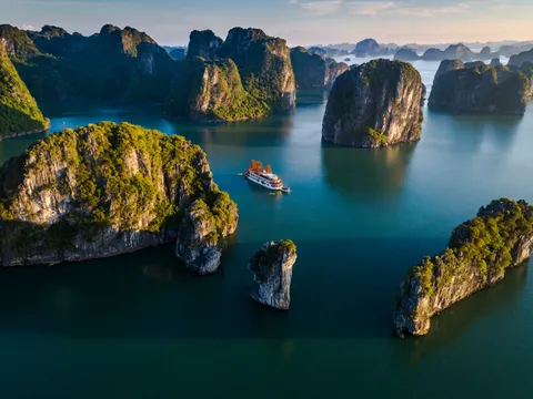 Aerial view of a cruise boat with red sails navigating between tall limestone karst islands in calm turquoise water.