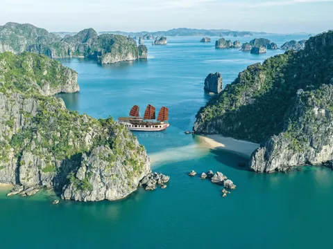 Aerial view of a white junk-style boat with three red-brown sails in turquoise water between green limestone karst islands, with small sandy beaches in the foreground.