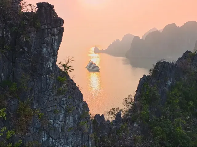 A cruise ship sails through calm water between dramatic limestone karst formations at sunset or sunrise.