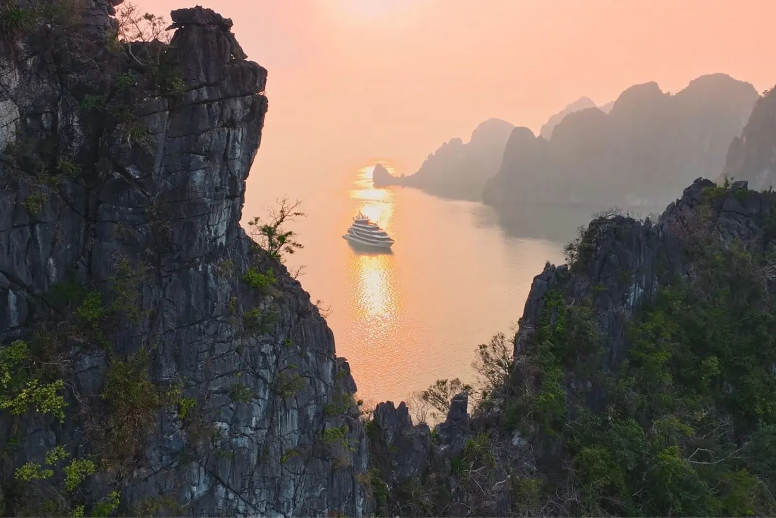 A cruise ship sails through calm water between dramatic limestone karst formations at sunset or sunrise.