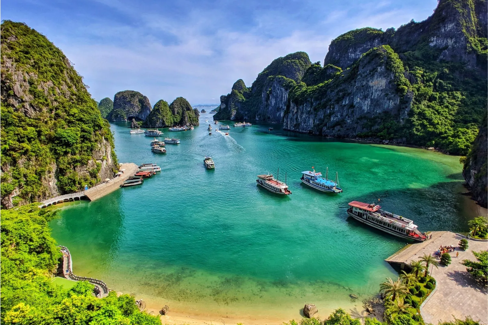 An aerial view of a sheltered bay with turquoise water, limestone karst cliffs covered in vegetation, several cruise boats moored near wooden docks, and a small beach.
