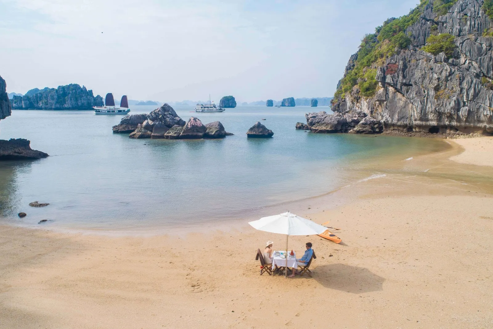 Two people dining at a white-clothed table under a white umbrella on a sandy beach, with karst limestone formations and cruise boats visible in the calm bay behind them.