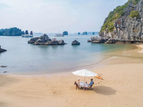 Two people dining at a white-clothed table under a white umbrella on a sandy beach, with karst limestone formations and cruise boats visible in the calm bay behind them.