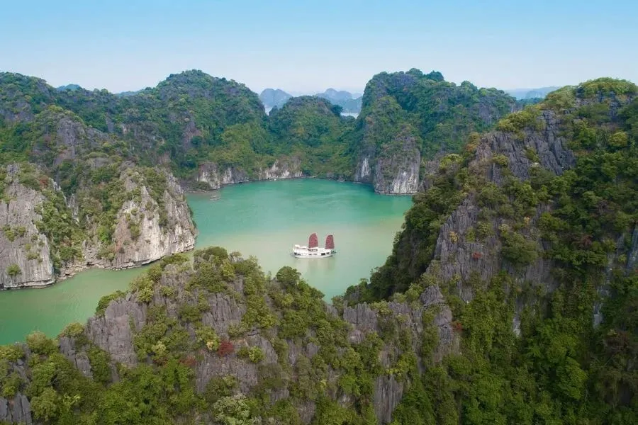 An aerial view of a white boat with two red sails on turquoise water, enclosed by steep limestone karst cliffs covered in green vegetation.