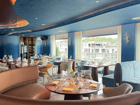 A curved blue-ceilinged restaurant interior with set tables and tan and blue seating, and a window showing a docked white cruise ship with a green hill behind it; a logo is visible on the neighbouring ship's hull.