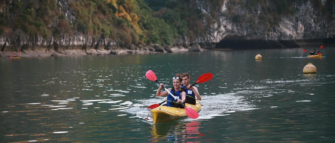 Two people kayaking in a yellow kayak with red paddles on calm water surrounded by limestone karst cliffs.