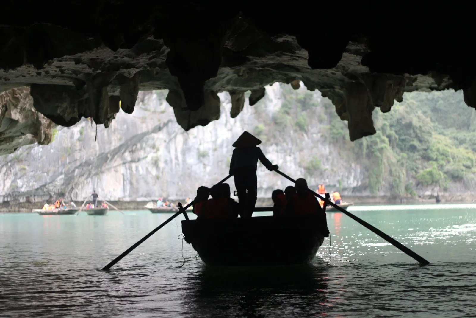 A silhouetted rower in a conical hat steers a small rowing boat carrying passengers in orange life jackets out from under a limestone cave overhang, with other small boats and karst cliffs beyond.