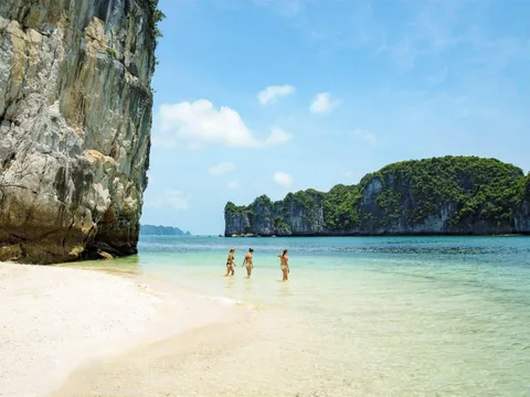 Three people in swimwear stand in shallow turquoise water near a white sand beach with dramatic limestone karst cliffs in the background.