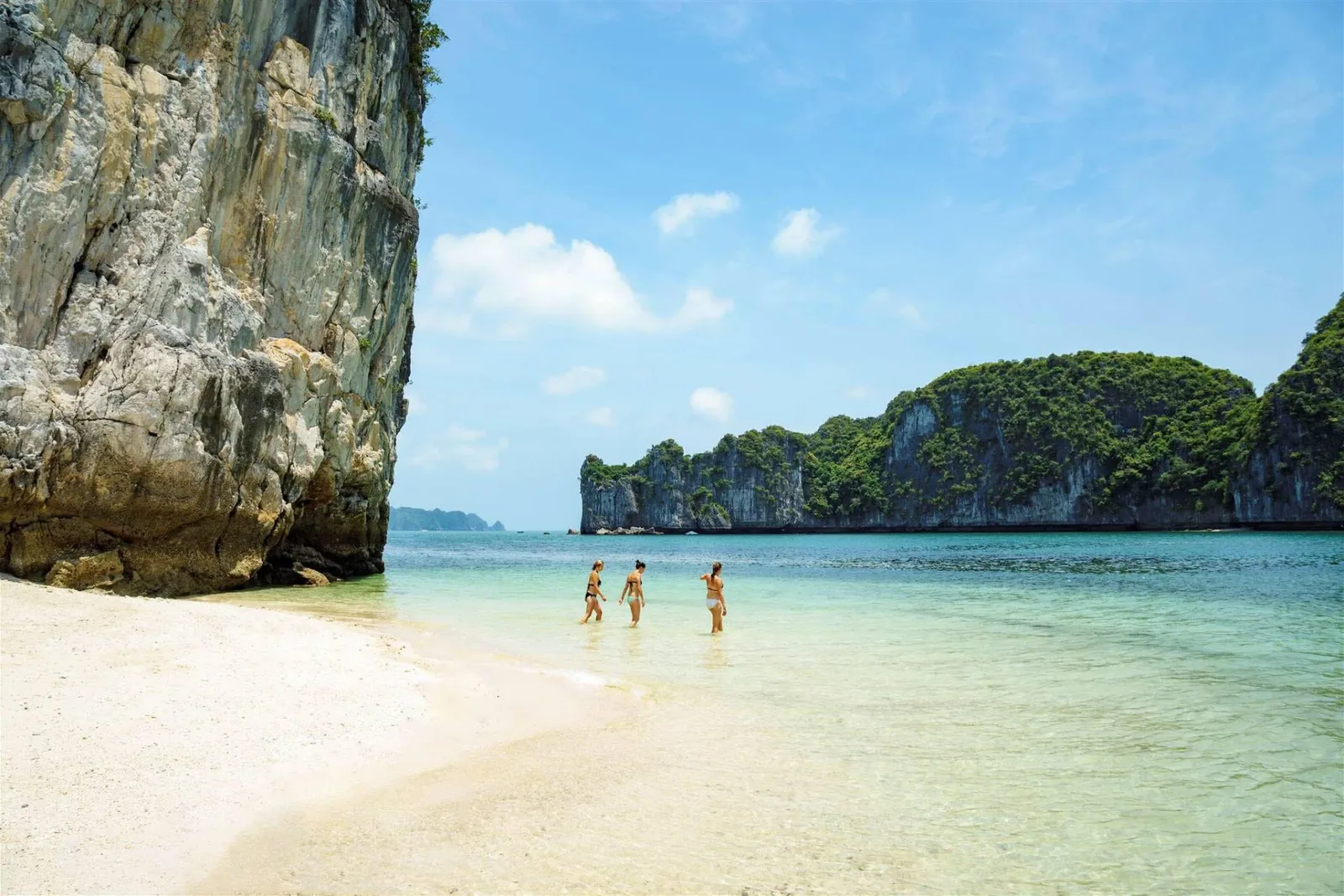 Three people in swimwear stand in shallow turquoise water near a white sand beach with dramatic limestone karst cliffs in the background.
