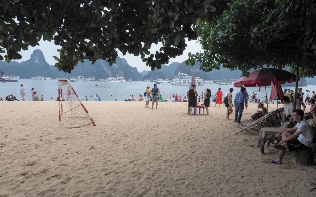A beach with people, umbrellas, and volleyball net with limestone karst islands and cruise boats visible across the water.