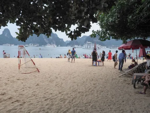 A beach with people, umbrellas, and volleyball net with limestone karst islands and cruise boats visible across the water.