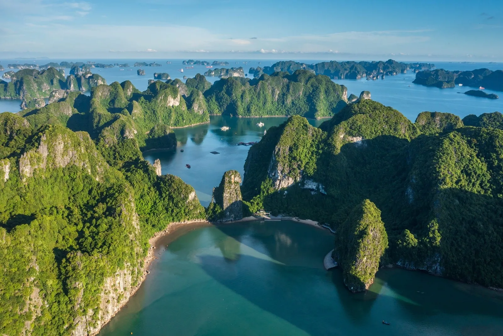 Aerial view of green forested limestone karst islands surrounding a turquoise bay with a small beach and tiny distant boats.