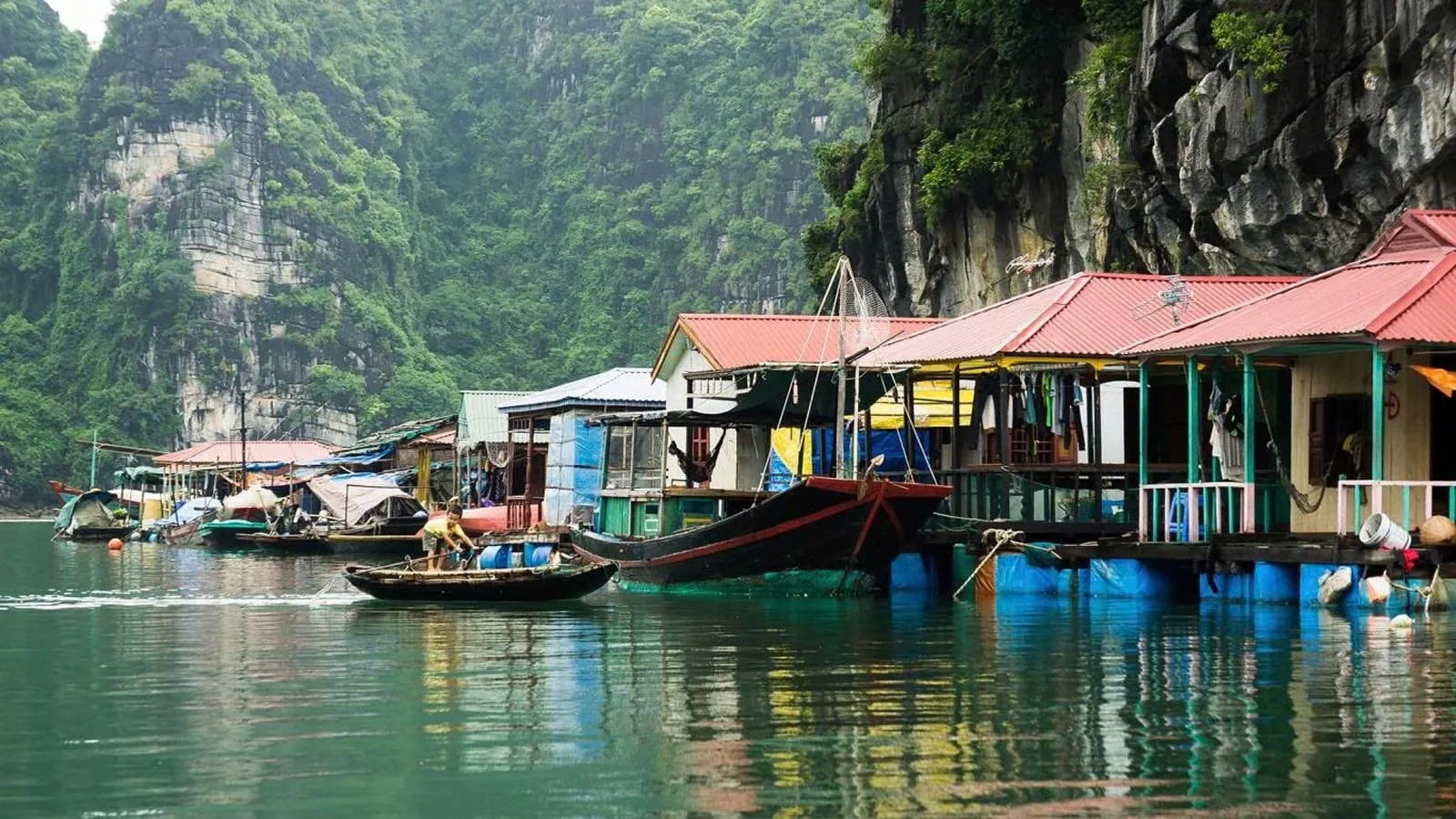 A floating village with houses on pontoons and small boats sits against dramatic limestone karst cliffs covered in vegetation.