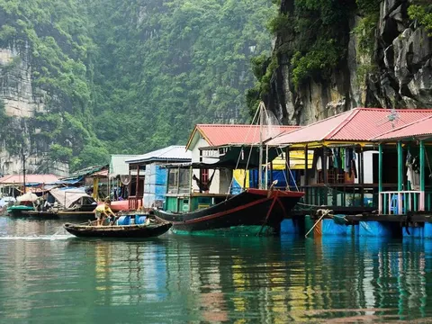 A floating village with houses on pontoons and small boats sits against dramatic limestone karst cliffs covered in vegetation.