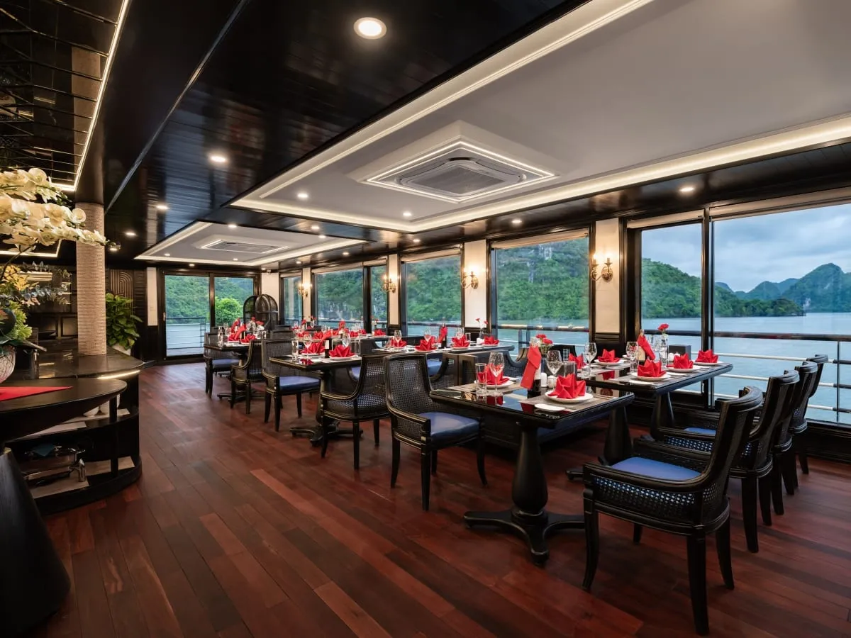 An indoor dining room on a boat with dark wood tables, black rattan chairs with blue cushions, and red folded napkins, with large windows showing green limestone karst islands and calm water.