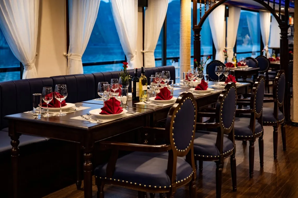 A dining room with a long dark wood table set with wine glasses, plates and red folded napkins, navy studded chairs and a banquette, and white curtains framing windows that show dusky water and dark karst-like hills.