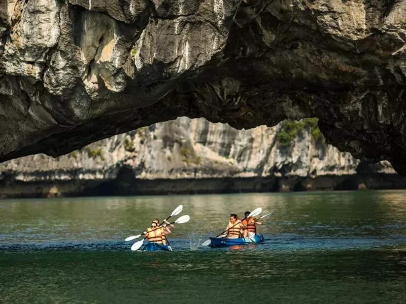 Two people in orange life jackets kayaking through a cave opening with limestone karst formations visible through the arch.