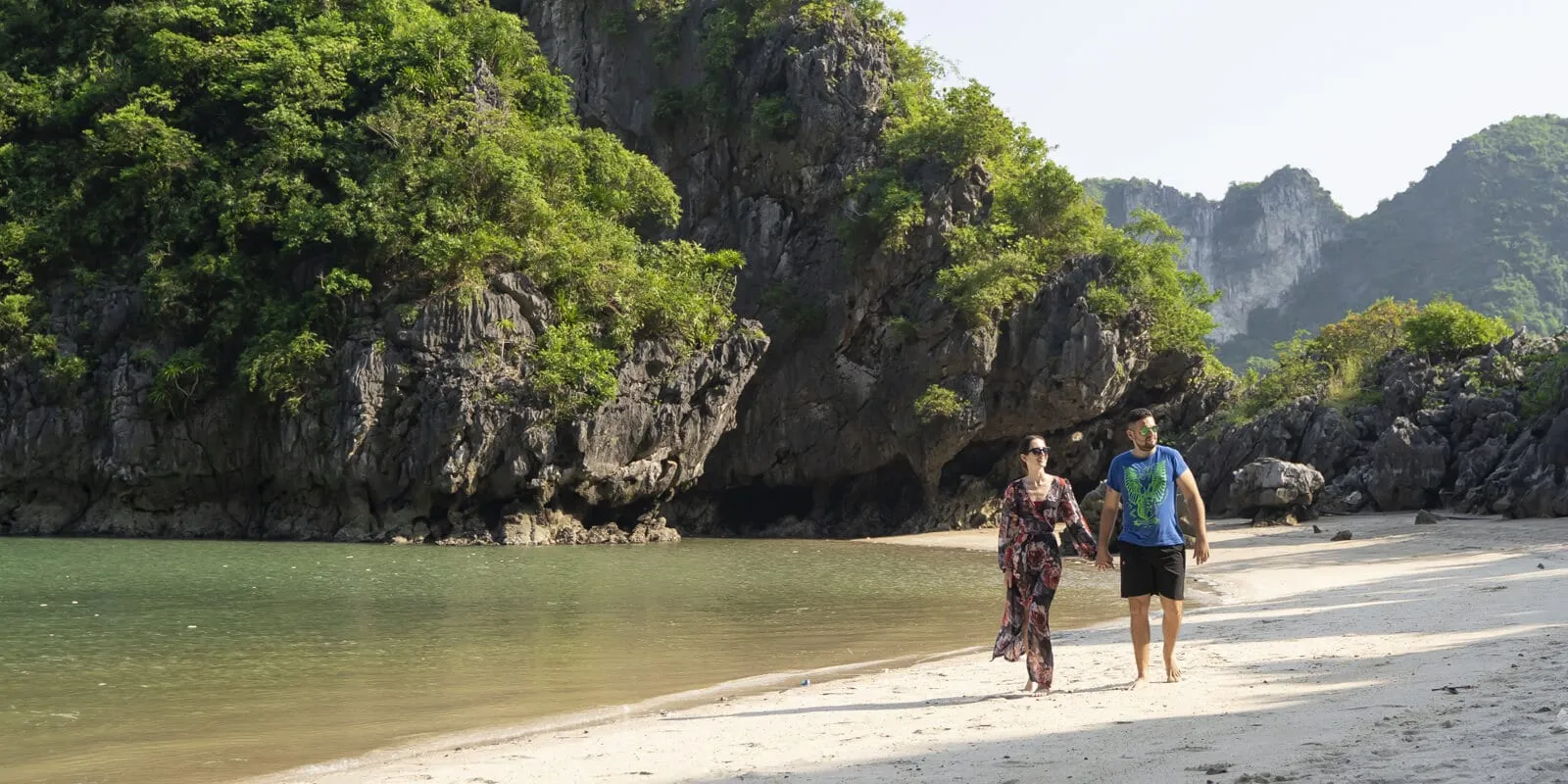 A man and woman walk hand in hand along a white sand beach beside green water, with dark limestone karst cliffs covered in vegetation behind them.